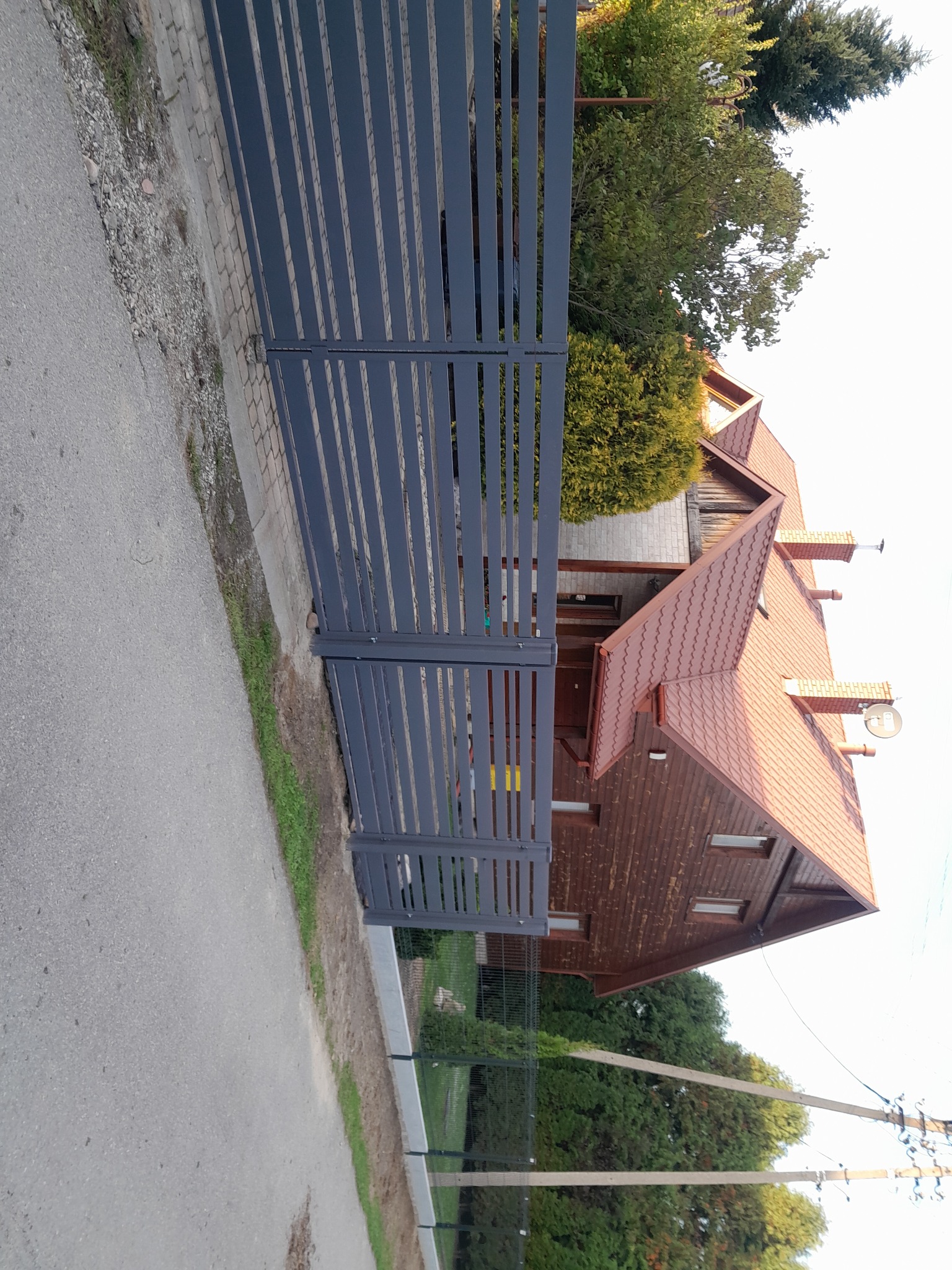 Nowoczesne, grafitowe ogrodzenie z poziomych paneli na tle drewnianego domu z czerwoną dachówką.