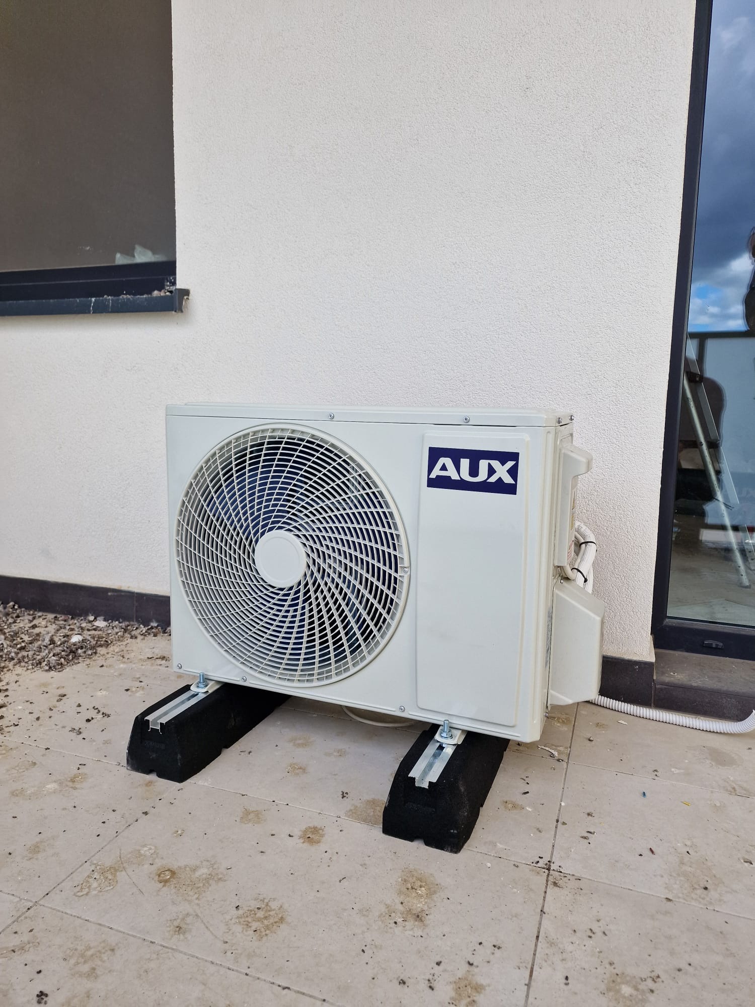 Zewnętrzna jednostka klimatyzacji marki AUX zamontowana na czarnych gumowych podkładkach, przy białej ścianie budynku z widocznym oknem i fragmentem drabiny.