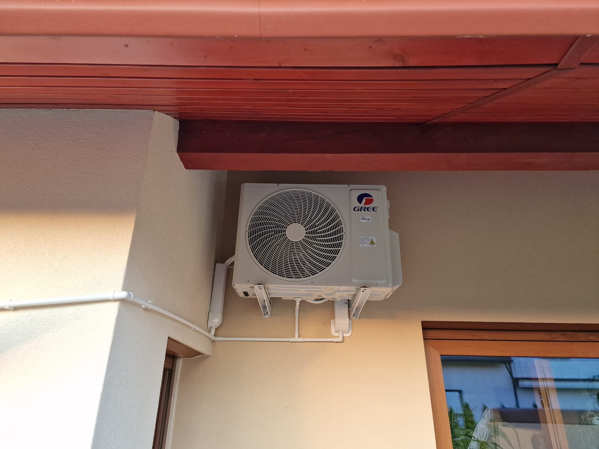 Zewnętrzna jednostka klimatyzacji marki Gree zamontowana na jasnej ścianie budynku, widoczne rury instalacyjne i drewniane elementy wykończenia dachu.