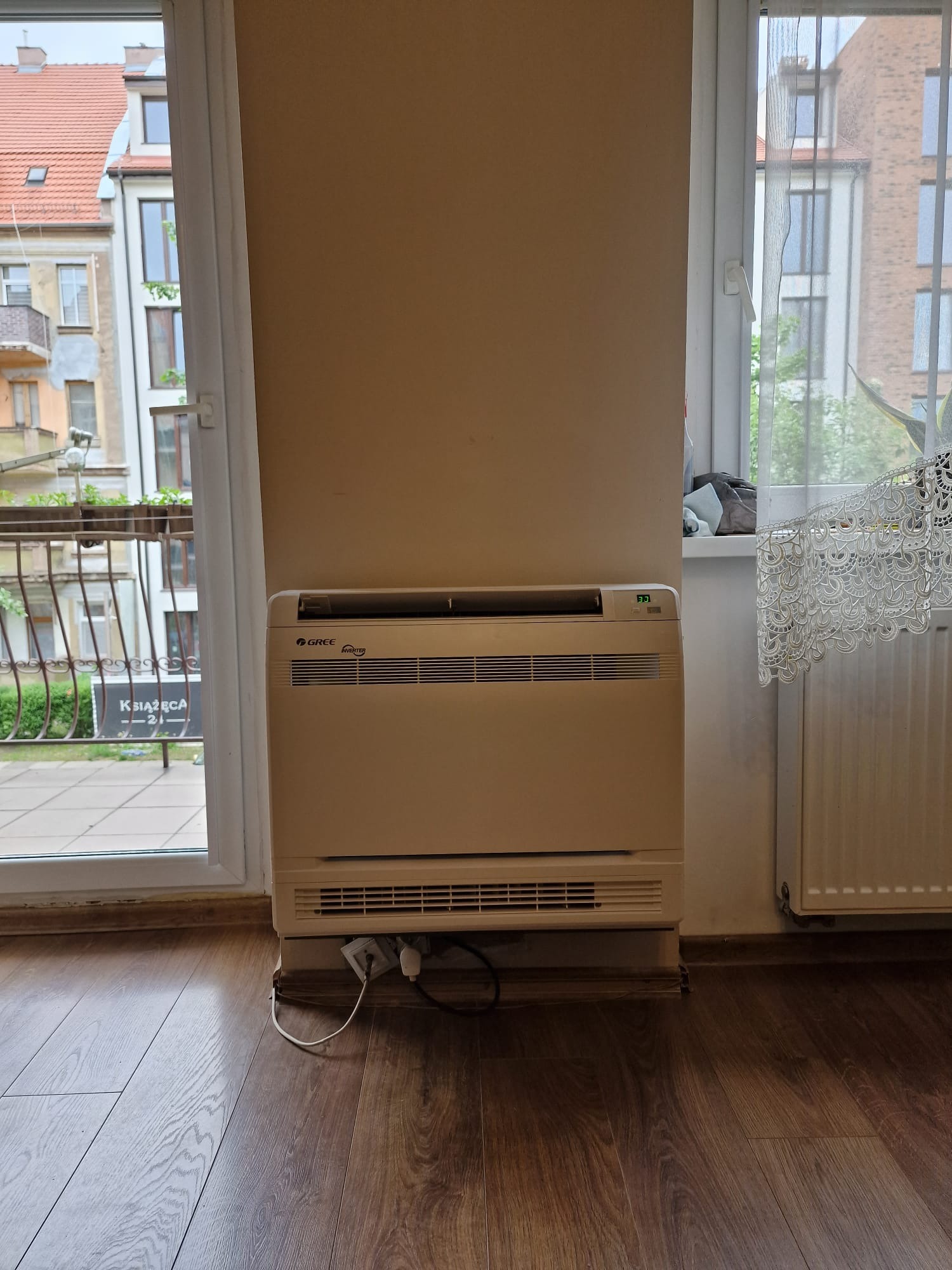 Stojący klimatyzator Gree Inverter w jasnym wnętrzu z widokiem na balkon i budynki mieszkalne, widoczne kable zasilające.