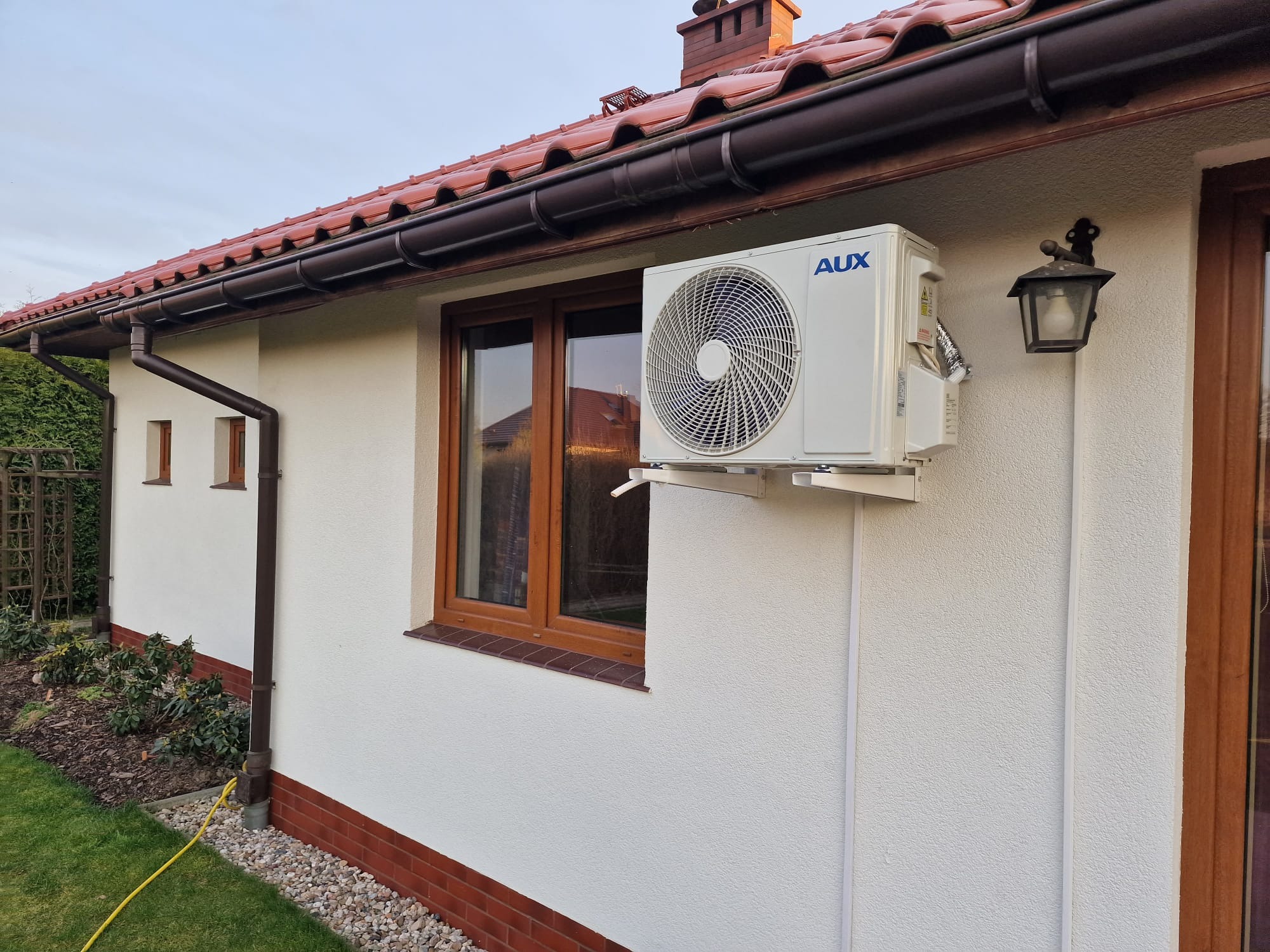 Biała jednostka zewnętrzna klimatyzatora AUX zamontowana na ścianie domu jednorodzinnego z brązową dachówką i oknami, widoczna lampa zewnętrzna i fragment ogrodu.