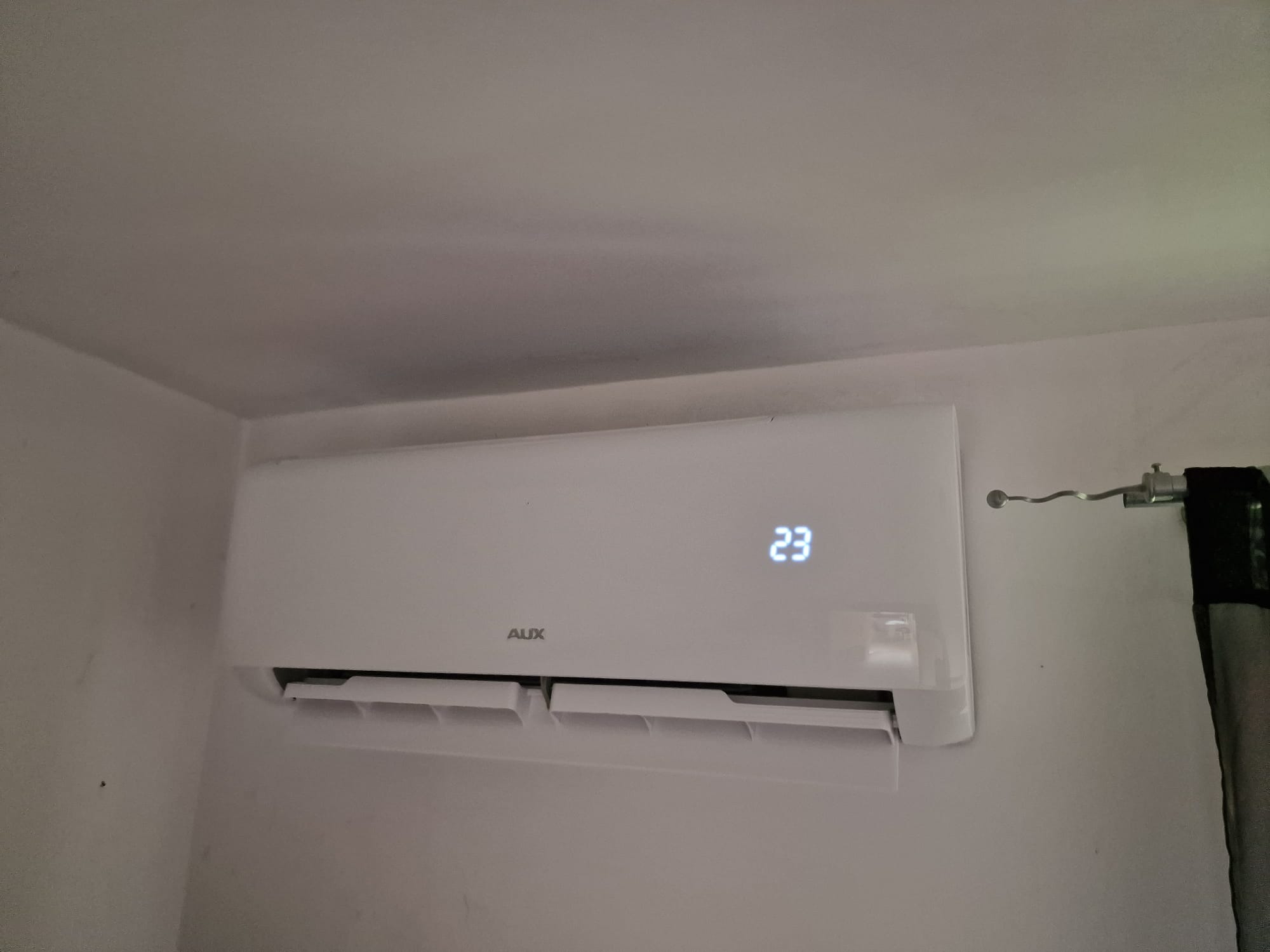 Biała jednostka klimatyzacji AUX zamontowana na ścianie, wyświetlająca temperaturę 23 stopnie, z widoczną zasłoną w tle.