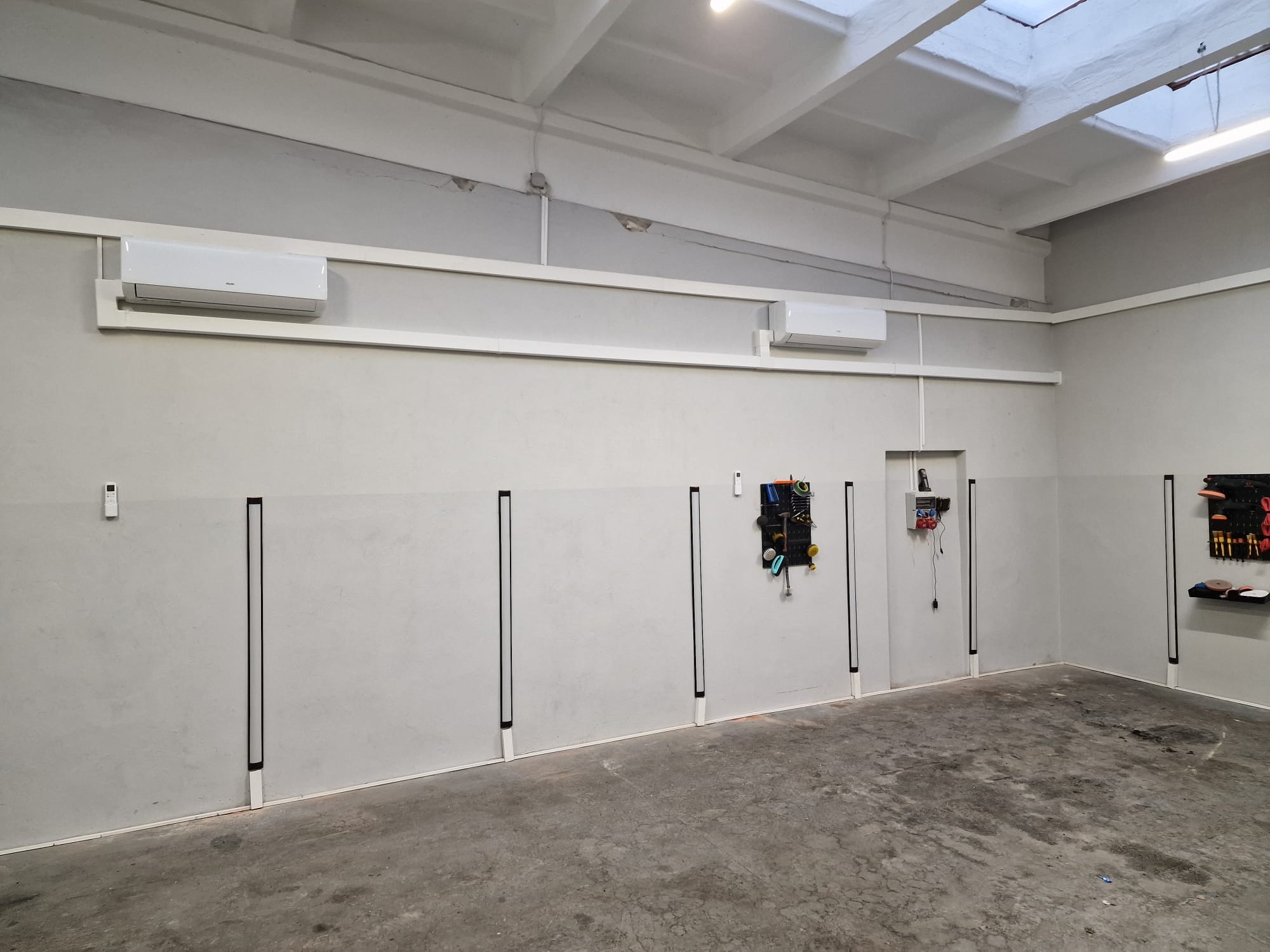 Wnętrze pomieszczenia z dwoma zamontowanymi klimatyzatorami ściennymi, widoczne narzędzia na ścianie, betonowa podłoga i białe ściany.