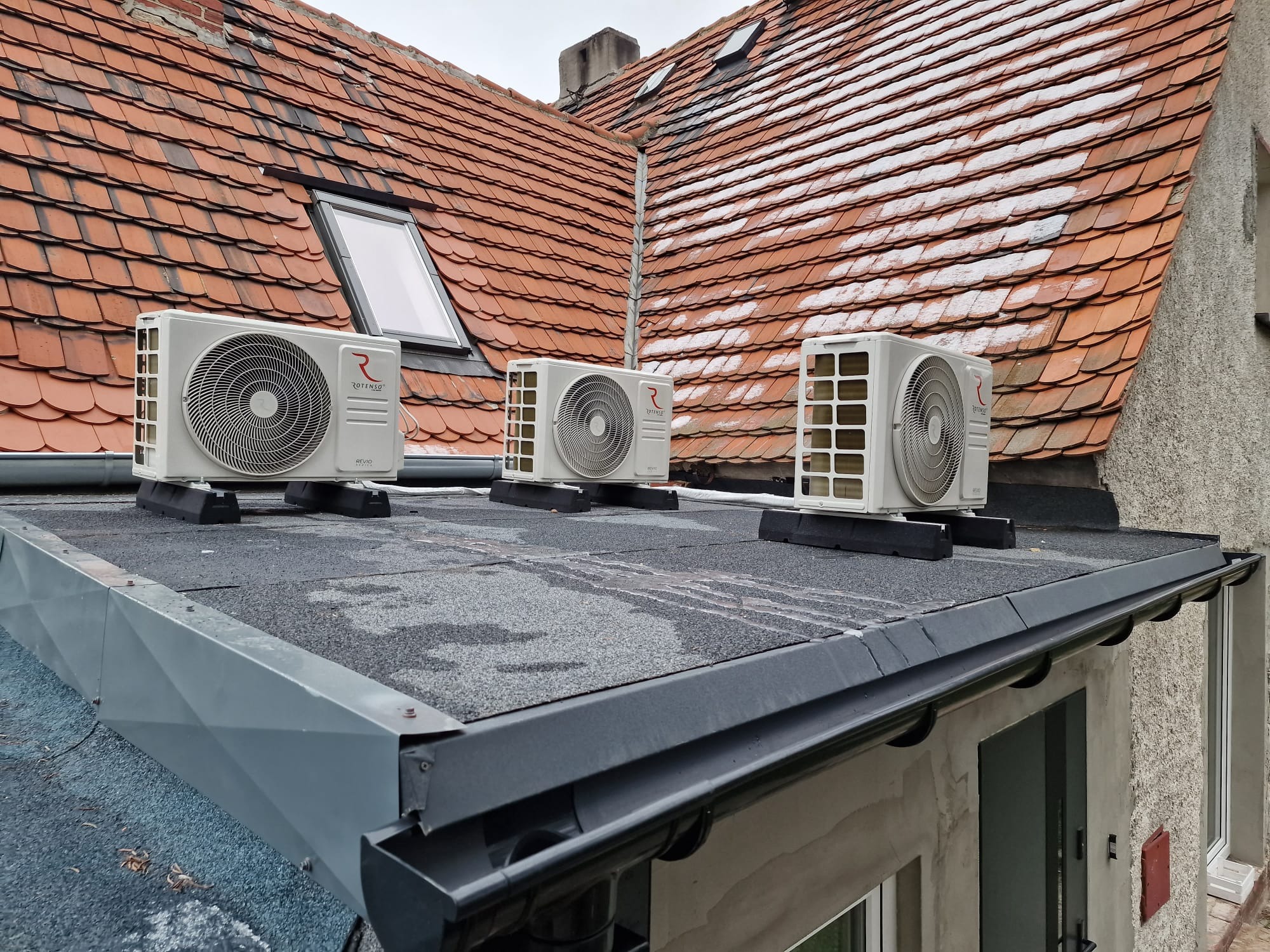 Trzy zewnętrzne jednostki klimatyzacji marki Rotenso zamontowane na płaskim dachu budynku z czerwoną dachówką w tle.