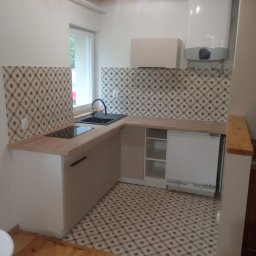 PrzemekKuchta - Nowoczesna, jasna kuchnia z drewnianym blatem, wzorzystą glazurą nad blatem i na podłodze, widok na zlew, płytę indukcyjną i bojler.