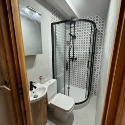 PrzemekKuchta - Mała łazienka po remoncie z kabiną prysznicową z czarną ramą, umywalką z szafką z jasnego drewna i toaletą, wykończona płytkami w biało-czarny wzór krzyżyków.