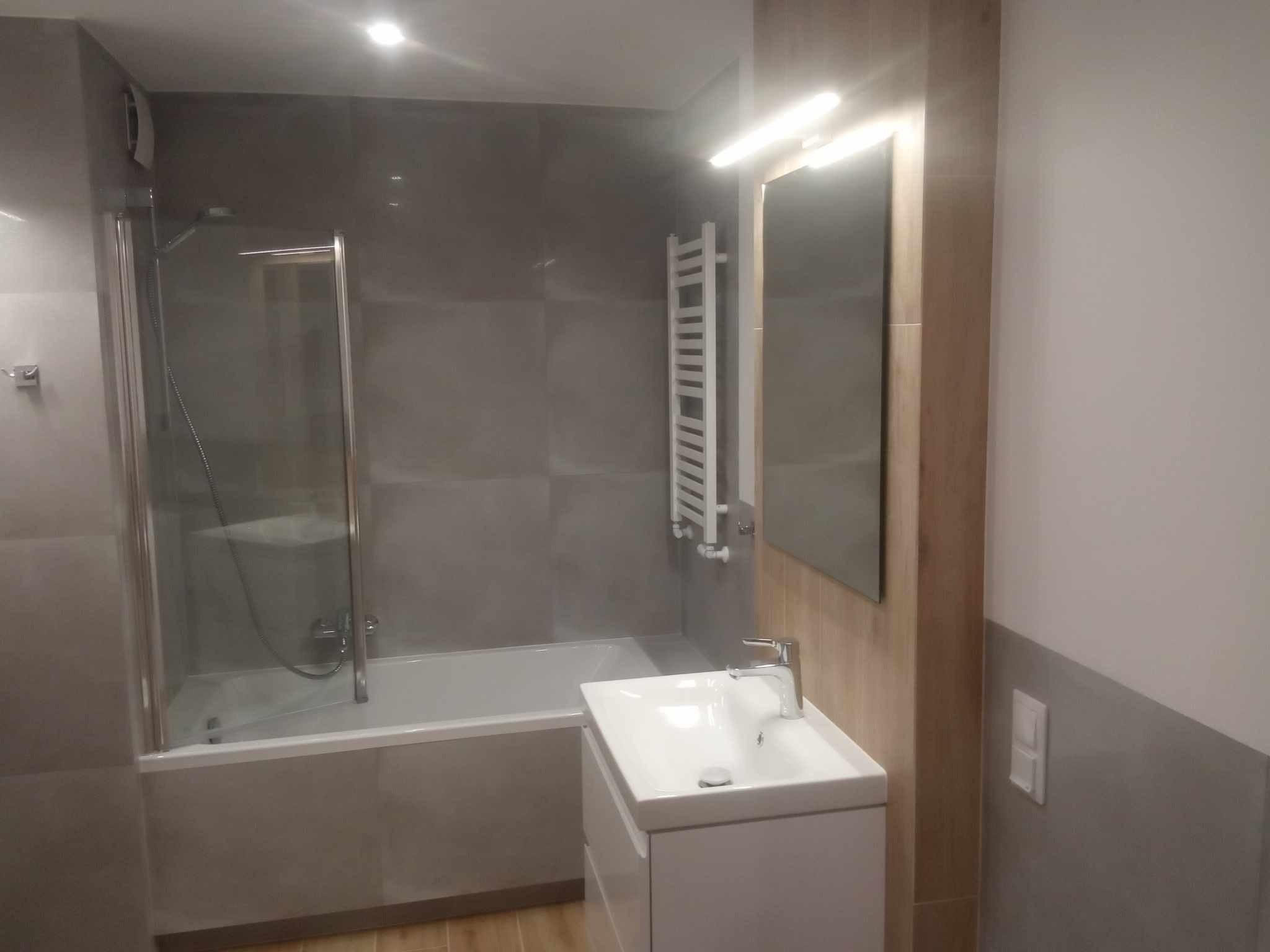 Nowoczesna łazienka z wanną za szklaną ścianką, umywalką nablatową na białej szafce i grzejnikiem drabinkowym na szarych płytkach ściennych. Lustro w drewnianej ramie i oświetlenie punktowe.