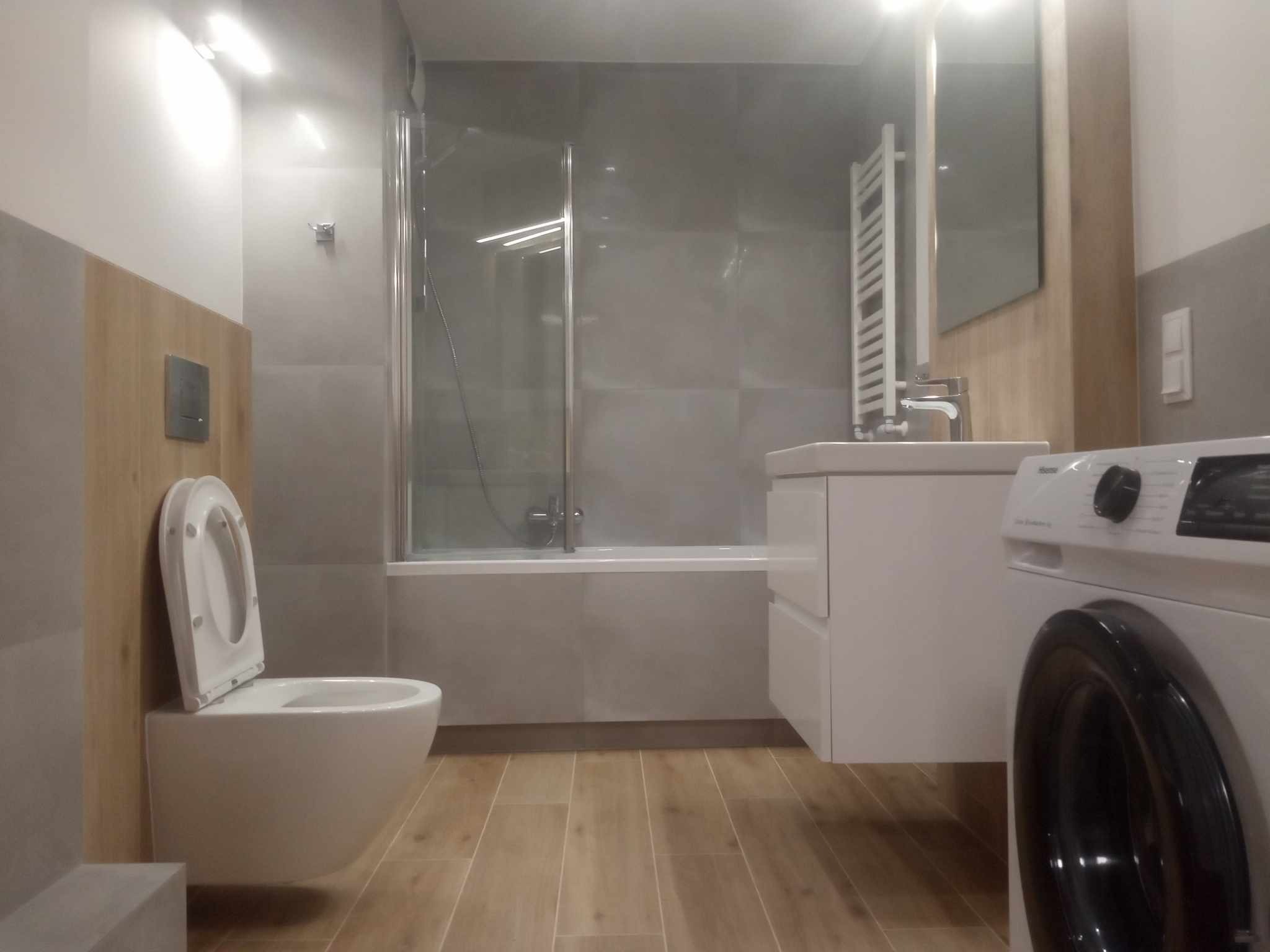 Nowoczesna łazienka z drewnopodobną podłogą, szarą glazurą, wiszącą toaletą z otwartą klapą, kabiną prysznicową i pralką z przodu po prawej stronie.