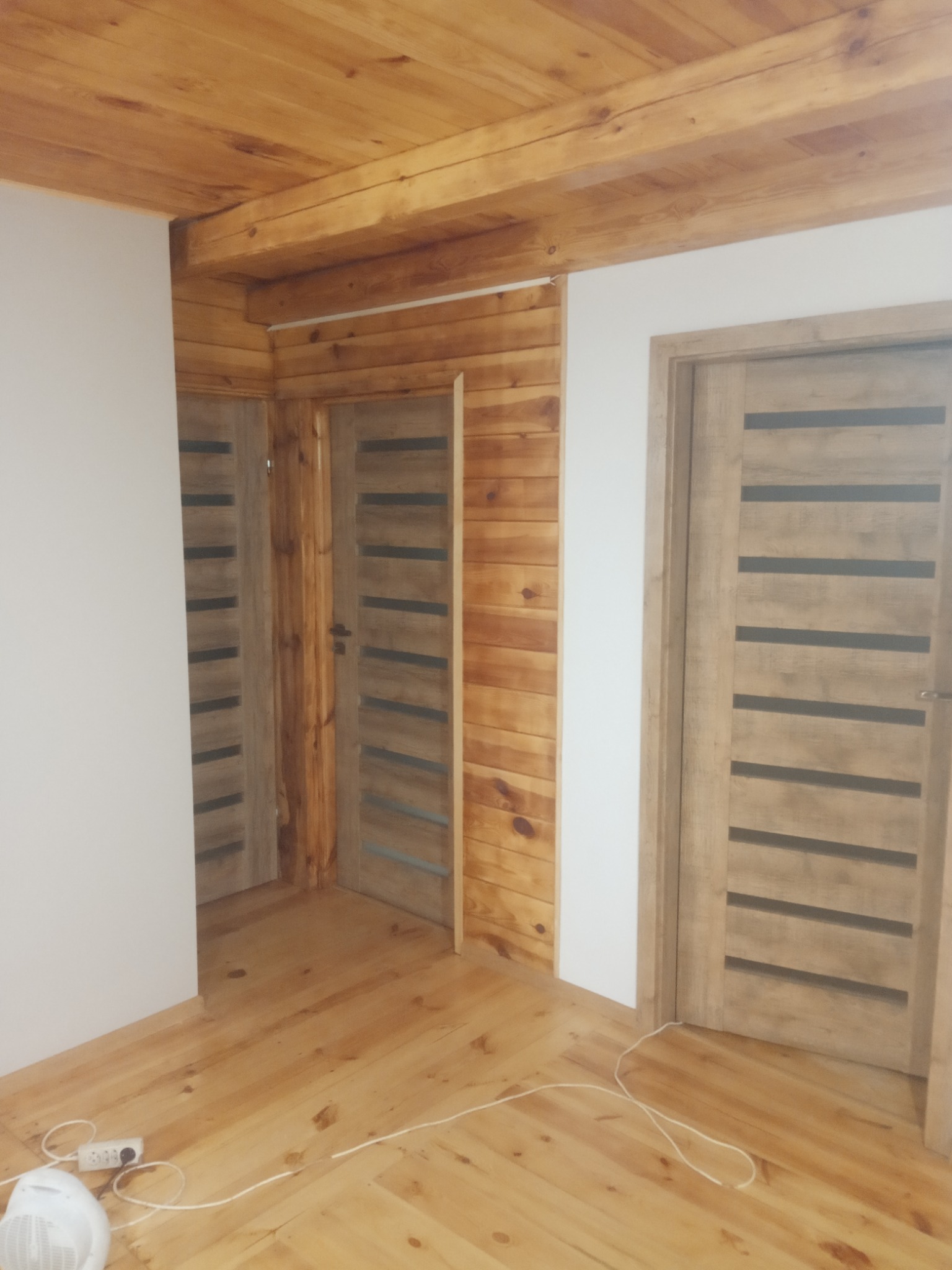 Wnętrze wykończone drewnem: sufit z belek, ściany obłożone boazerią, drewniana podłoga i trzy drewniane drzwi z poziomymi, ciemnymi paskami, widoczny kabel na podłodze.