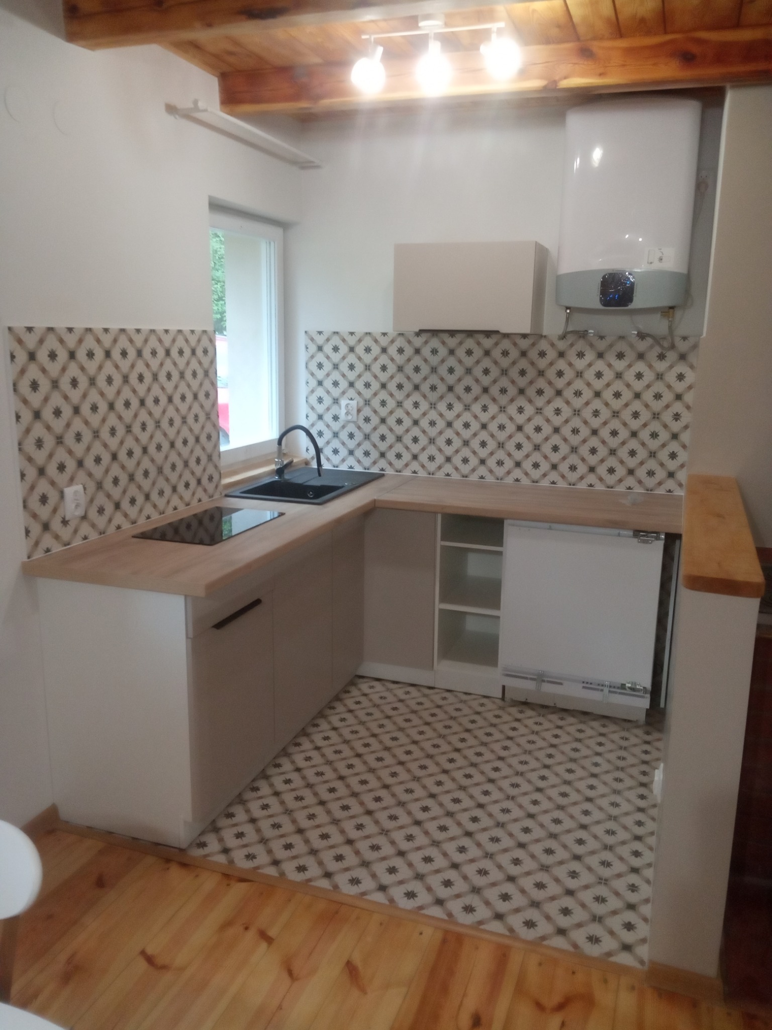 Nowoczesna, jasna kuchnia z drewnianym blatem, wzorzystą glazurą nad blatem i na podłodze, widok na zlew, płytę indukcyjną i bojler.