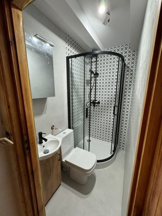 Mała łazienka po remoncie z kabiną prysznicową z czarną ramą, umywalką z szafką z jasnego drewna i toaletą, wykończona płytkami w biało-czarny wzór krzyżyków.