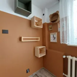 Ściana pomalowana na kolor terakoty z zamontowanymi drewnianymi półkami w kształcie sześcianów i prostokątów, powyżej linii farby biała ściana z półką w kształcie kosza misia, drewniana podłoga.