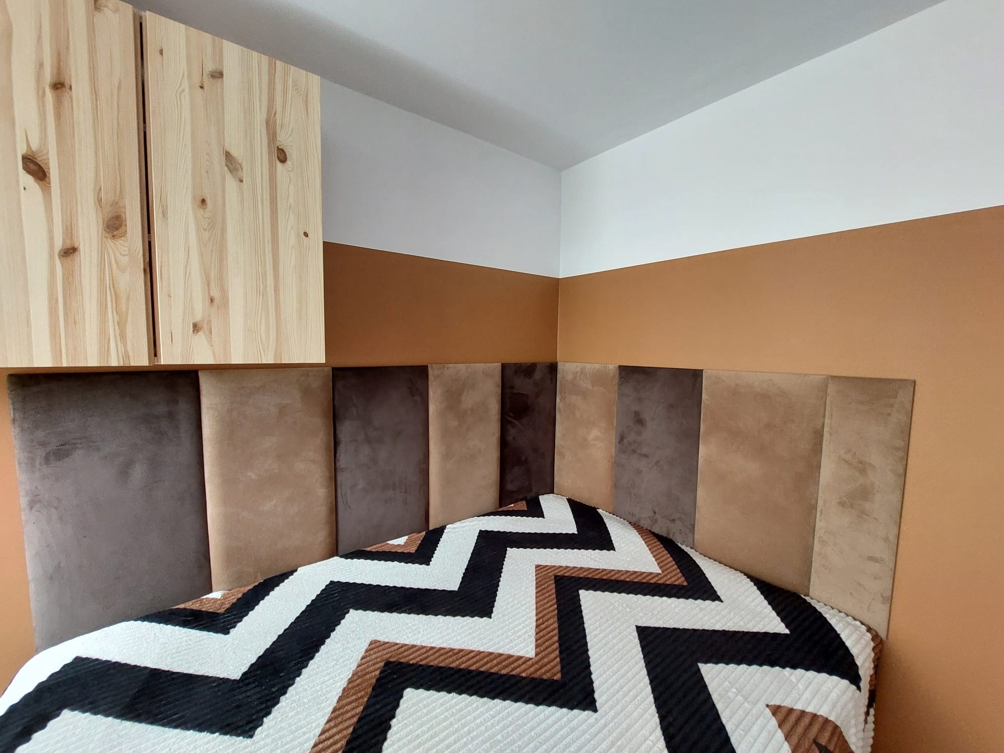 Sypialnia po odświeżeniu: ściana pomalowana dwukolorowo (biały i ciepły brąz), tapicerowany panelowy zagłówek w odcieniach beżu i brązu, drewniane szafki wiszące.