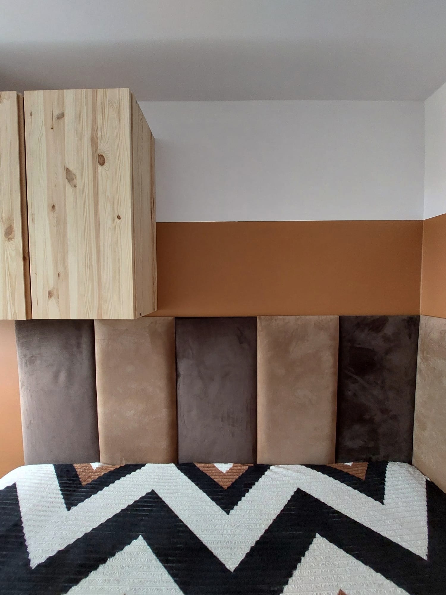 Panelowa ściana za łóżkiem w odcieniach brązu, nad nią pomalowana ściana w kolorze terakoty i biała góra, obok wiszą drewniane szafki.