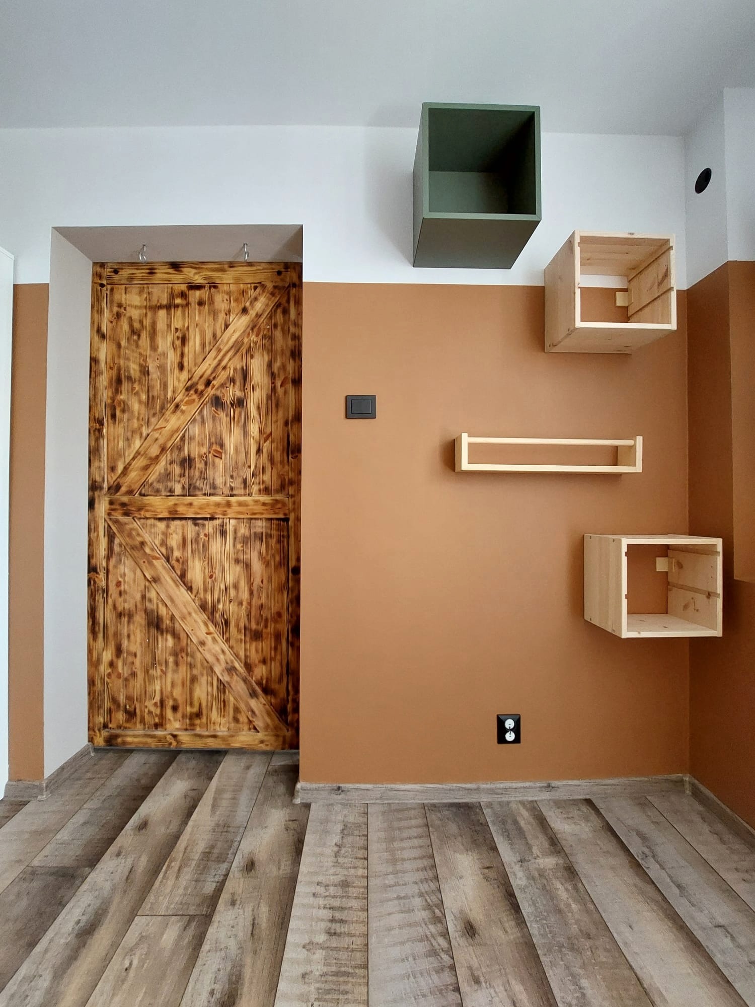 Wnętrze z drewnianą przesuwną bramą stodołową, panelami podłogowymi imitującymi drewno oraz półkami ściennymi w kształcie kostek i prostokąta na pomarańczowej ścianie.