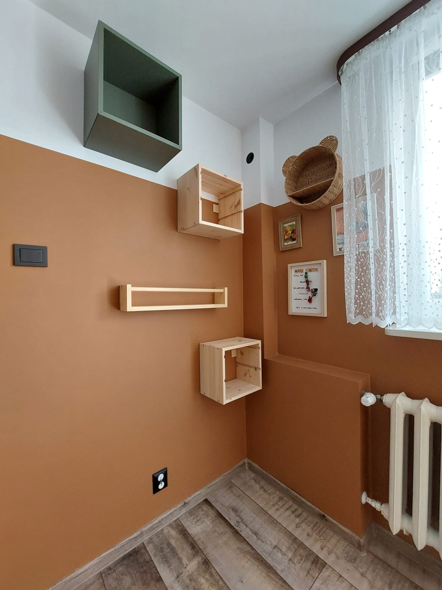 Ściana pomalowana na kolor terakoty z zamontowanymi drewnianymi półkami w kształcie sześcianów i prostokątów, powyżej linii farby biała ściana z półką w kształcie kosza misia, drewniana podłoga.
