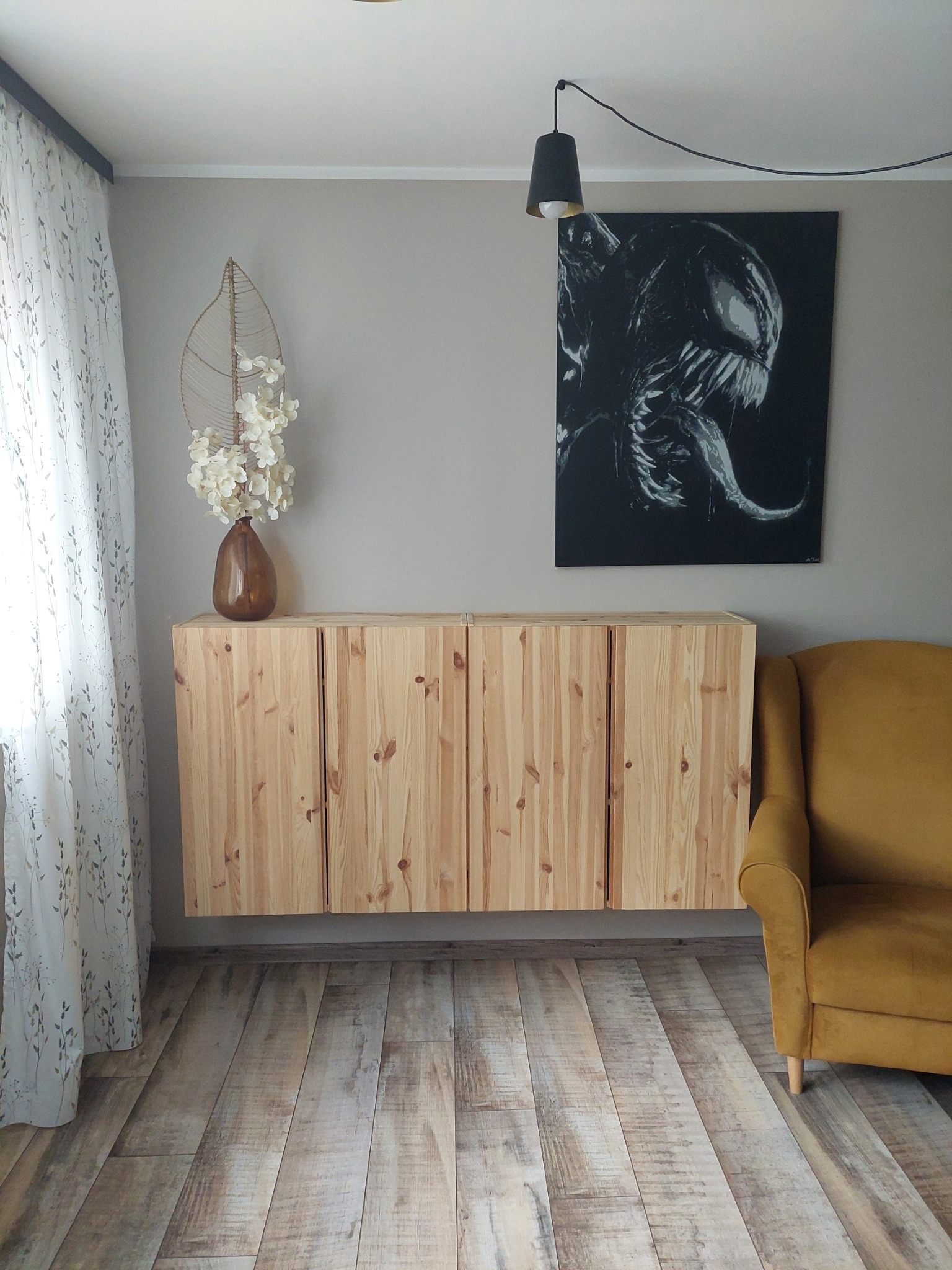 Nowoczesny salon z podwieszaną szafką z jasnego drewna, obrazem w czerni i bieli oraz musztardowym fotelem, uzupełniony dekoracjami i wzorzystą zasłoną.