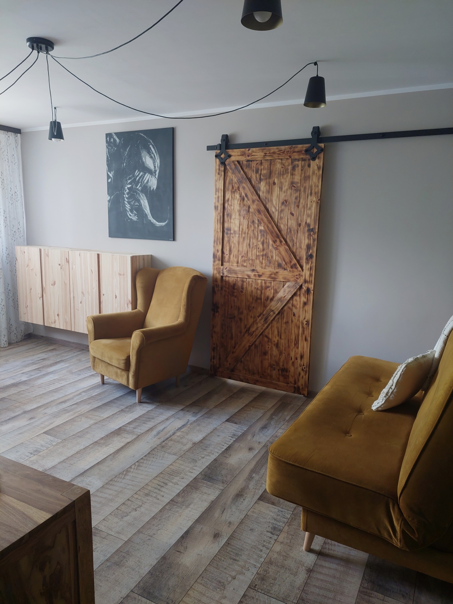 Nowoczesny salon z drewnianą przesuwną bramą stodołową, musztardowym fotelem i sofą, oraz podłogą imitującą naturalne drewno.