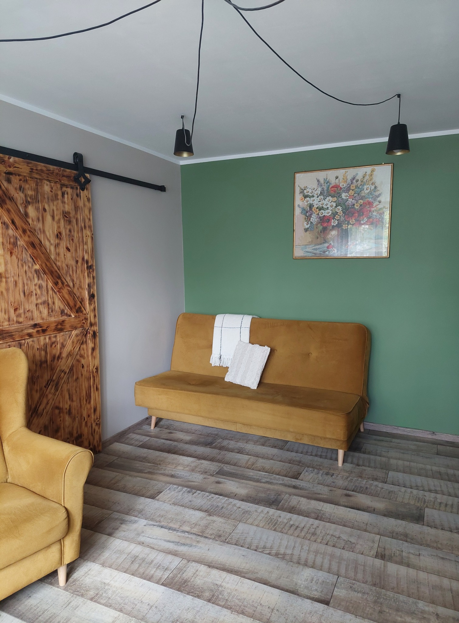 Rustykalne wnętrze z drewnianymi drzwiami przesuwnymi, musztardową sofą z poduszkami i obrazem kwiatów na zielonej ścianie, uzupełnione przez lampy wiszące na kablach i podłogę imitującą drewno.