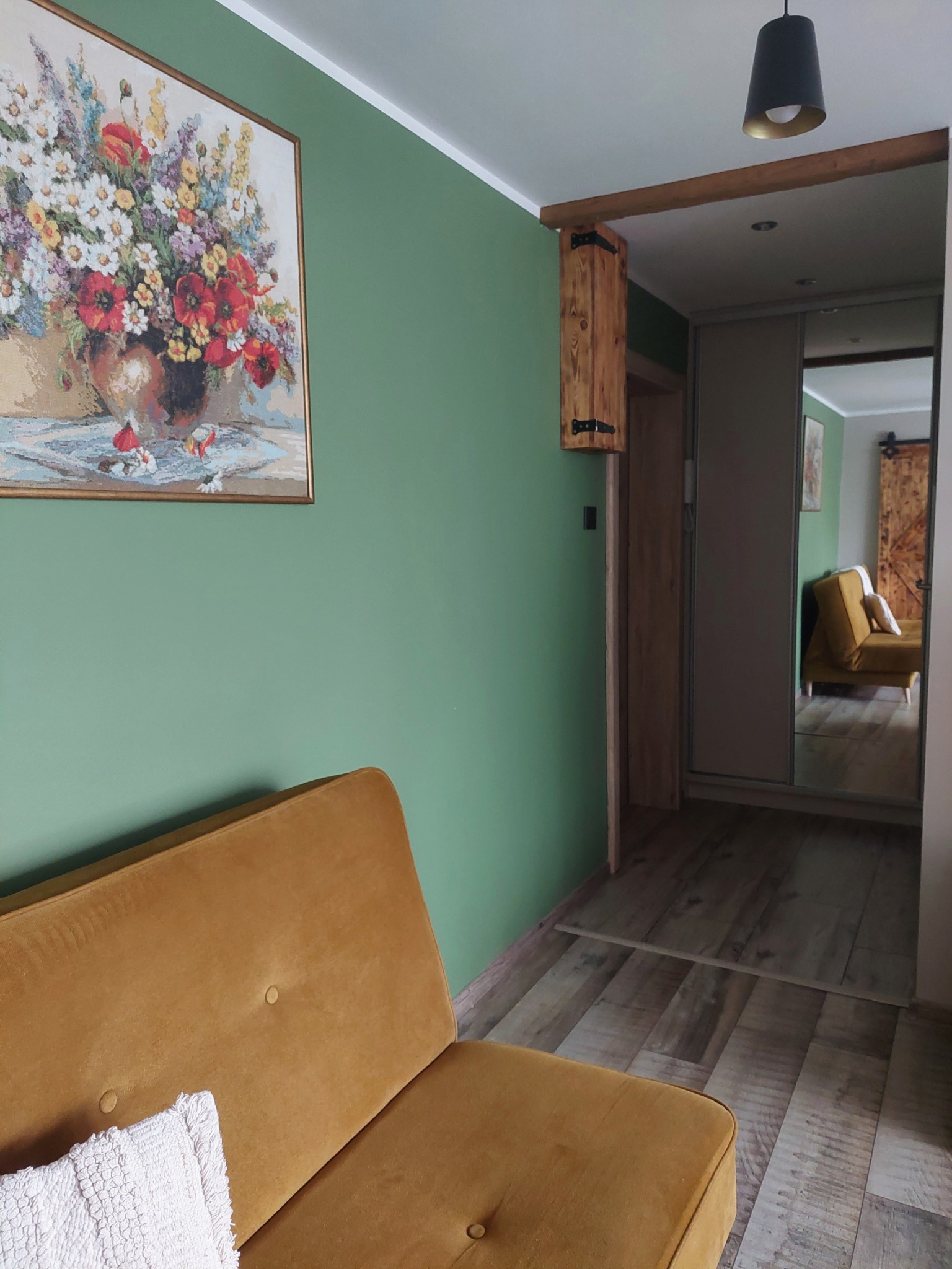 Wnętrze pokoju z zieloną ścianą, obrazem kwiatów, musztardową sofą i podłogą imitującą drewno, widoczne drzwi przesuwne z lustrem i drewniana szafka nad przejściem.
