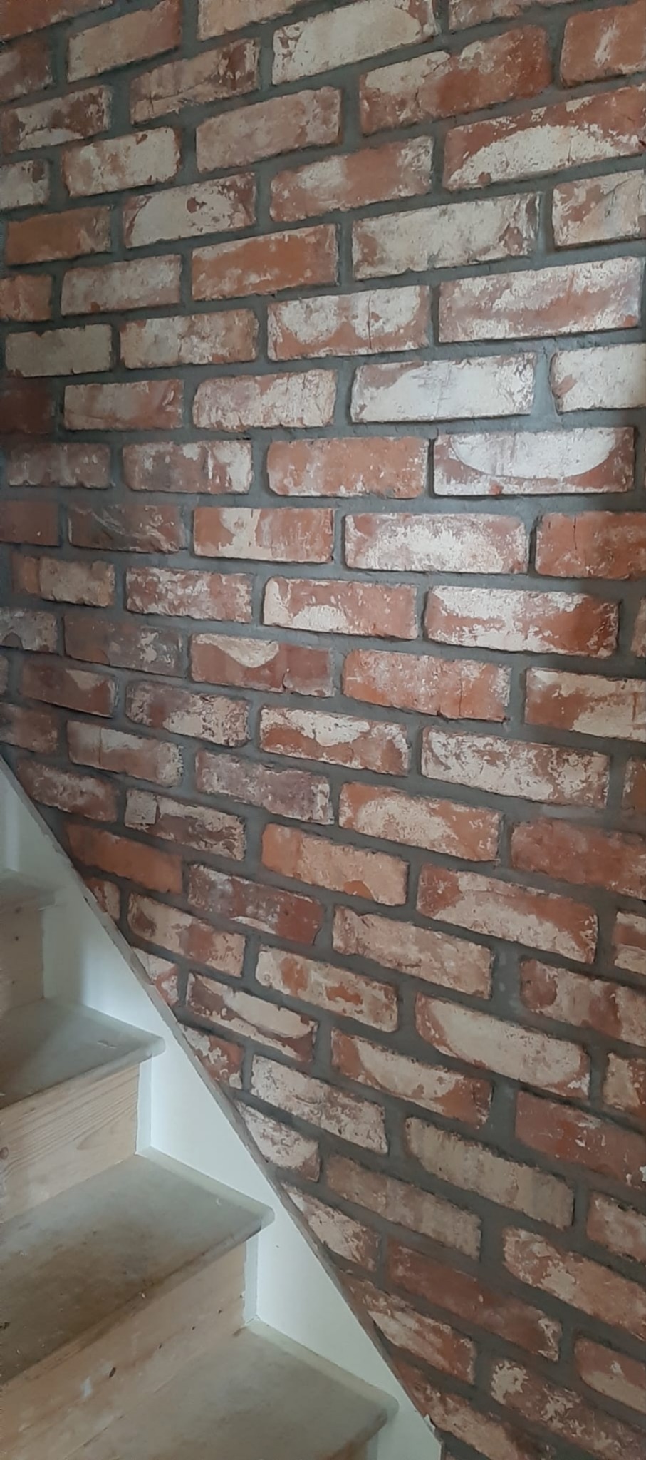 Ściana wykończona cegłą dekoracyjną w odcieniach czerwieni i bieli, przylegająca do drewnianych schodów z białymi listwami przypodłogowymi.