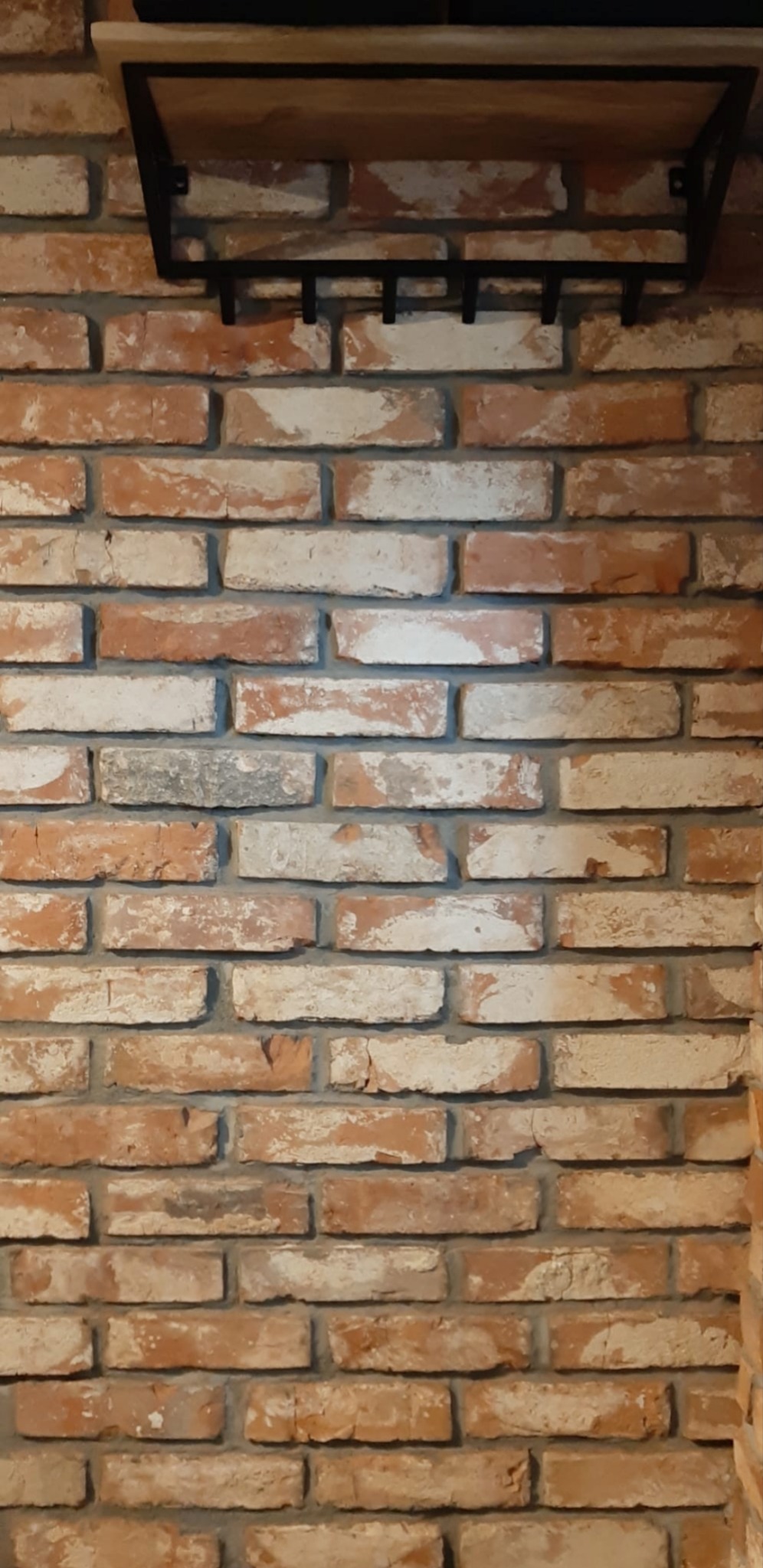 Ściana z cegły klinkierowej w odcieniach czerwieni i bieli, z półką z jasnego drewna i metalowym, czarnym wspornikiem z haczykami.