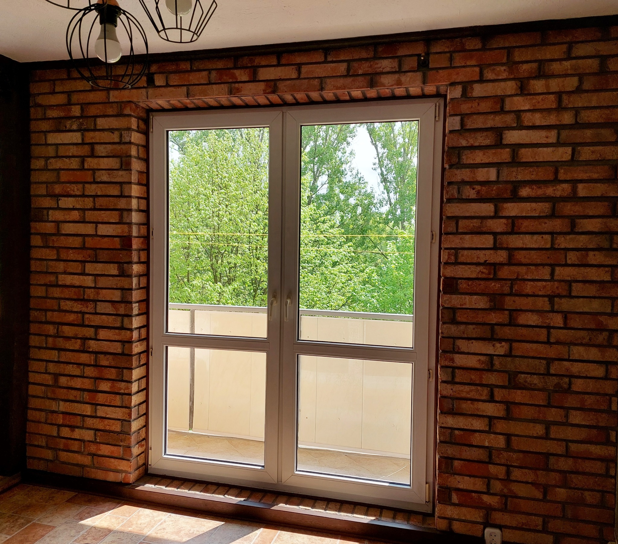 Nowe dwuskrzydłowe okno balkonowe z białymi ramami w ścianie z czerwonej cegły, widok na zieleń za oknem, oświetlenie sufitowe w stylu loft.