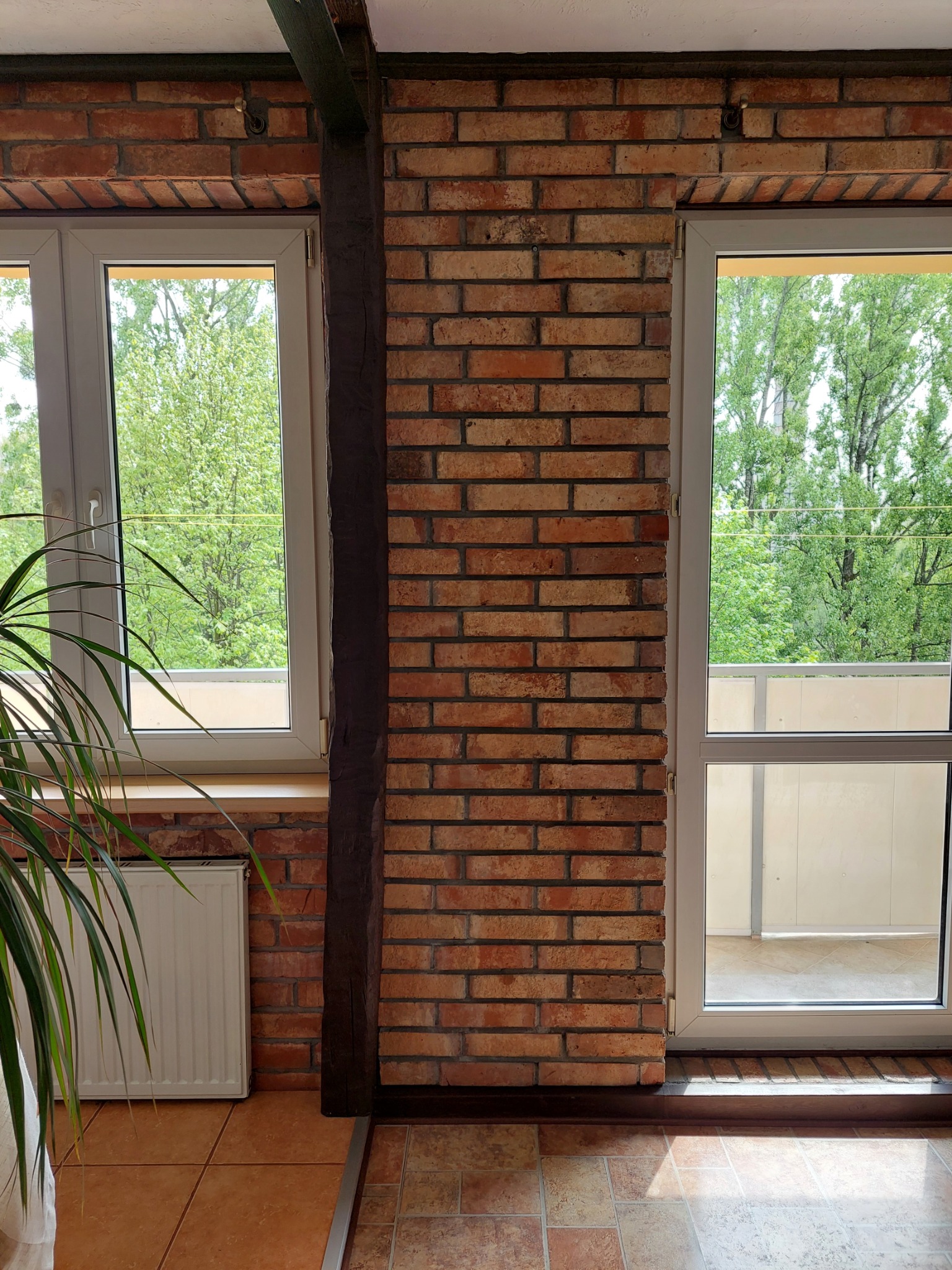Wnętrze z ceglaną ścianą, białymi oknami z widokiem na zieleń i drewnianą belką, na podłodze terakota.
