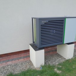 PGTECH SPÓŁKA Z OGRANICZONĄ ODPOWIEDZIALNOŚCIĄ - Zewnętrzna jednostka pompy ciepła Heitztechnik Callaverde, zamontowana na betonowych postumentach przy ścianie budynku, otoczona kostką brukową i kamieniami.