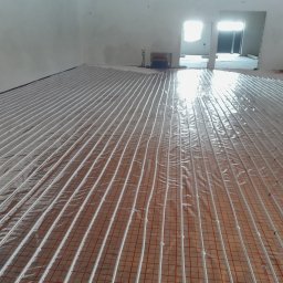 PGTECH SPÓŁKA Z OGRANICZONĄ ODPOWIEDZIALNOŚCIĄ - Rozłożone rury ogrzewania podłogowego na folii z siatką, widoczny rozdzielacz w tle, surowy stan wykończenia wnętrza.