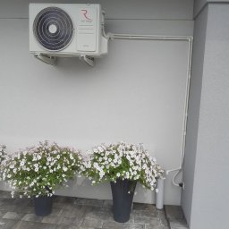 PGTECH SPÓŁKA Z OGRANICZONĄ ODPOWIEDZIALNOŚCIĄ - Zewnętrzna jednostka klimatyzacji marki Rotenso zamontowana na szarej ścianie budynku, z widocznymi rurami instalacyjnymi i dwoma donicami z białymi kwiatami na brukowanym chodniku.