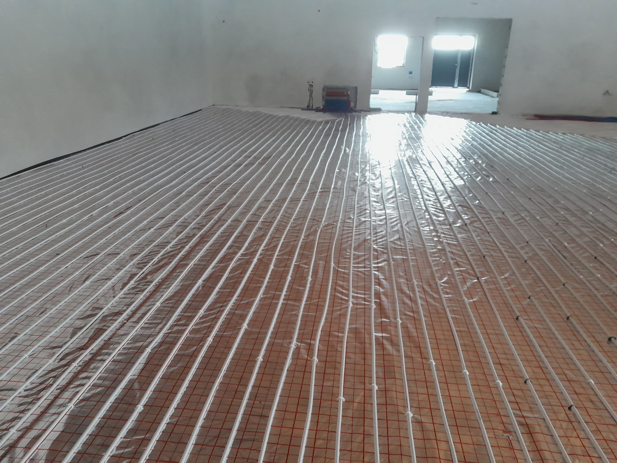 Rozłożone rury ogrzewania podłogowego na folii z siatką, widoczny rozdzielacz w tle, surowy stan wykończenia wnętrza.