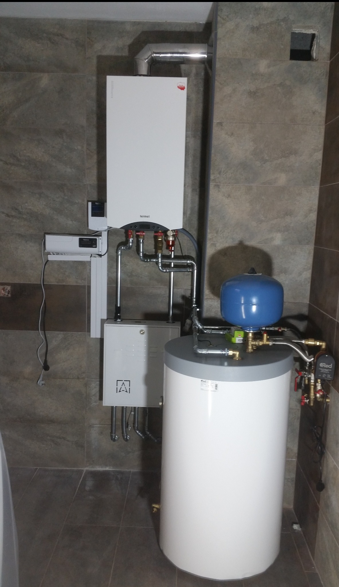 Nowoczesna kotłownia gazowa z automatyką, zawierająca biały piec Termet z odprowadzeniem spalin, zasobnik ciepłej wody z niebieskim naczyniem przeponowym oraz szafkę sterowniczą z logo w kształcie...