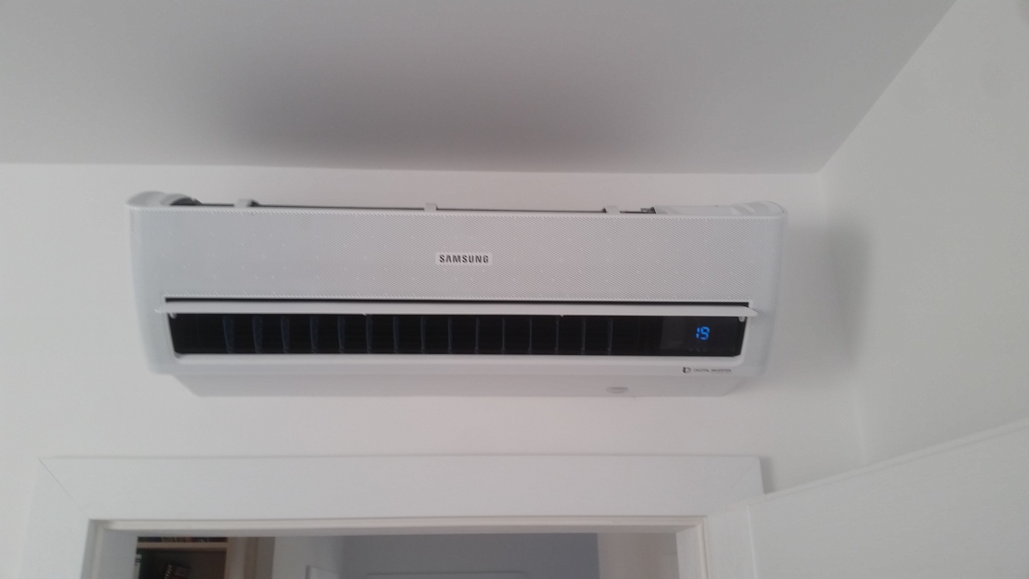 Biała jednostka wewnętrzna klimatyzatora Samsung zamontowana na ścianie nad ościeżnicą drzwi, wyświetlająca temperaturę 19 stopni Celsjusza.