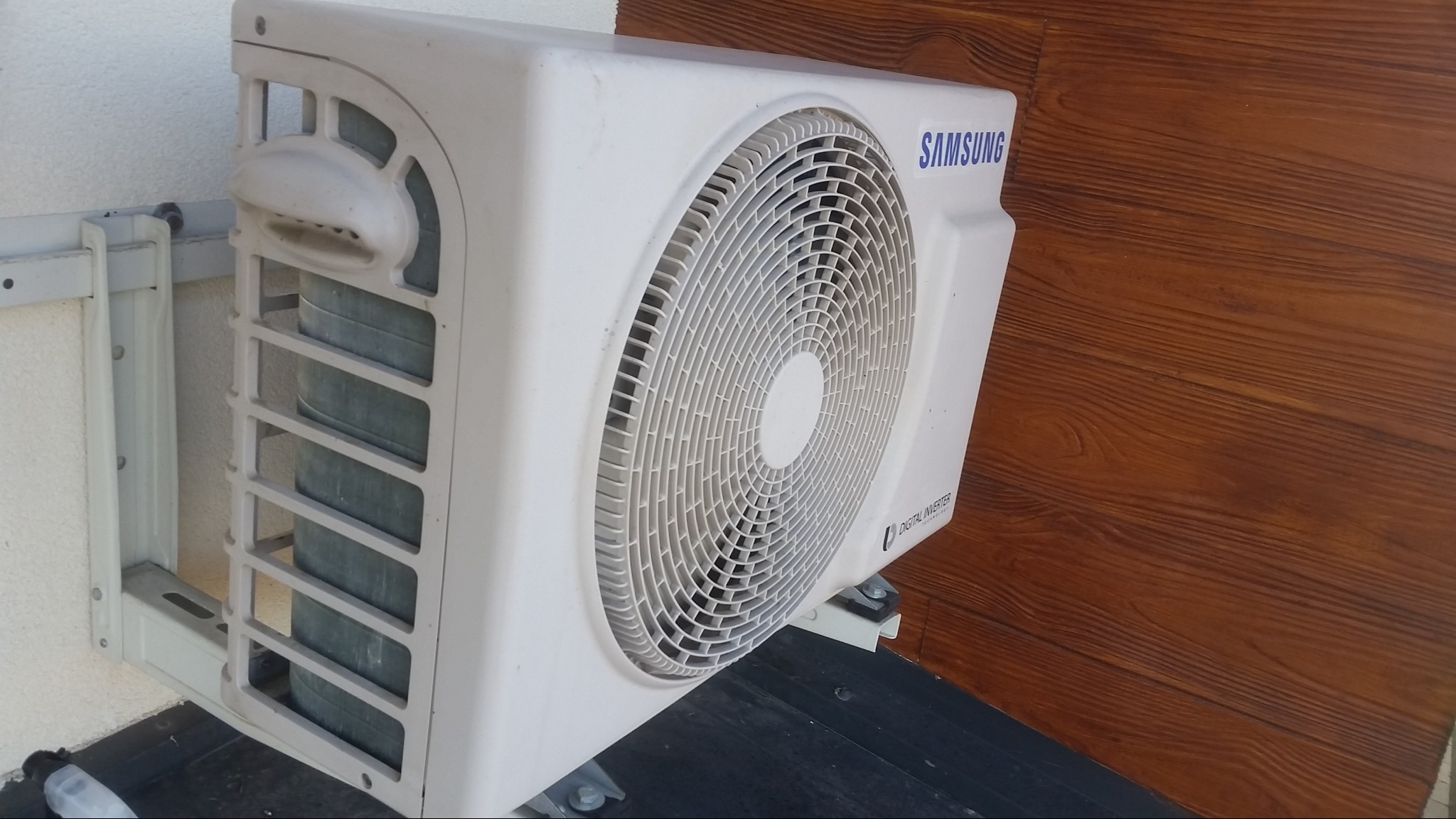 Zewnętrzna jednostka klimatyzatora Samsung zamontowana na ścianie budynku z drewnianym wykończeniem.
