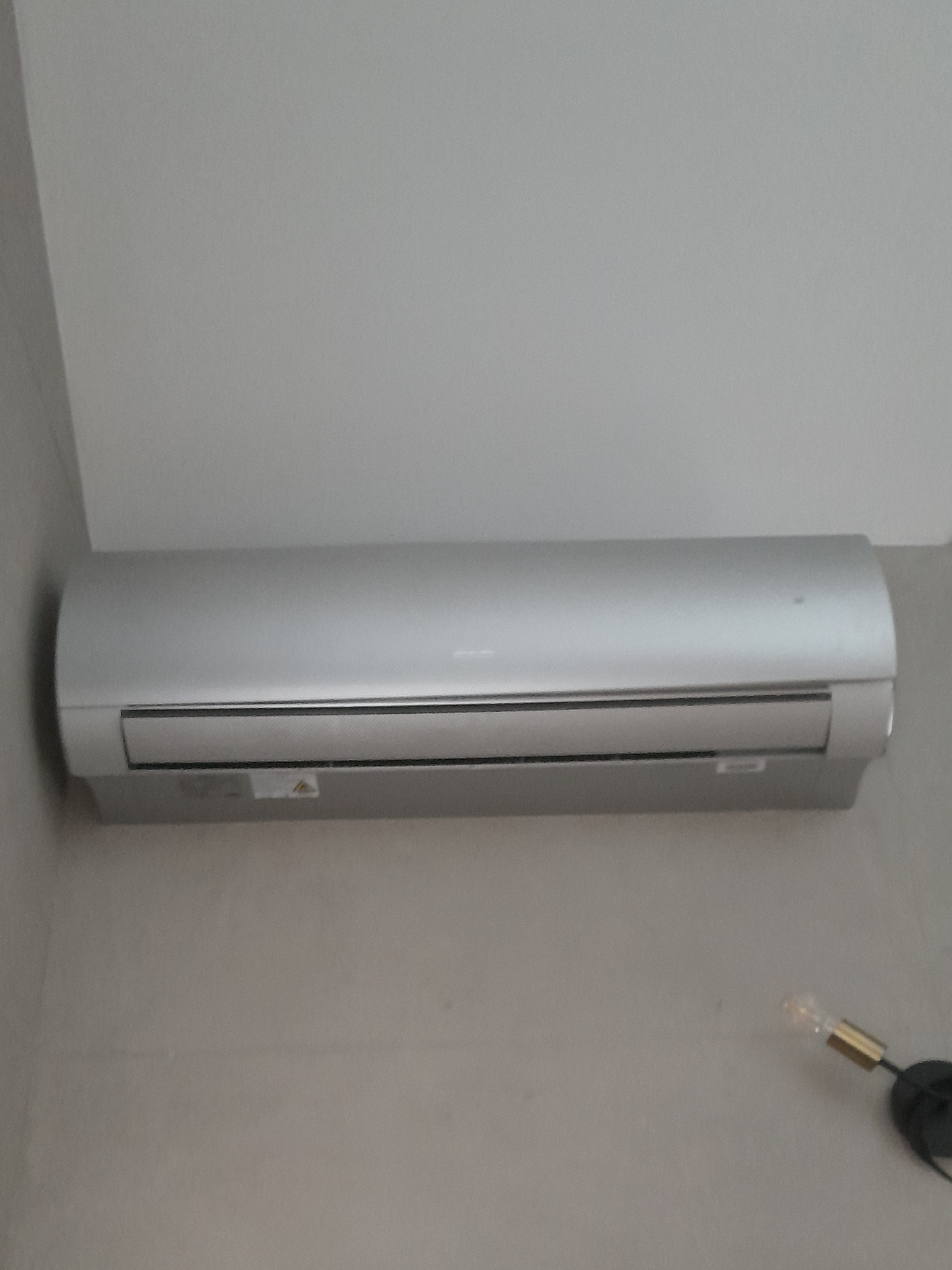 Szary klimatyzator ścienny zamontowany w pomieszczeniu, widoczny fragment lampy na ścianie.