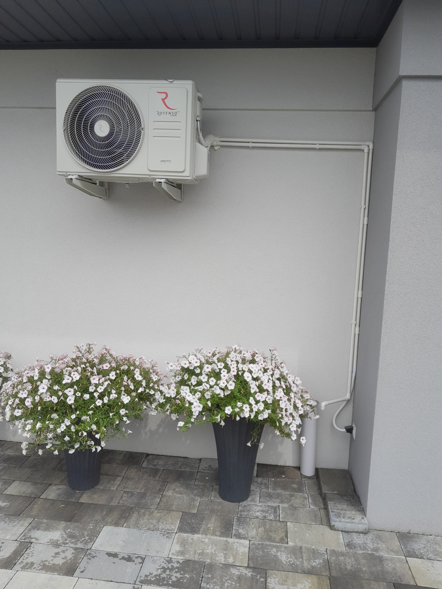 Zewnętrzna jednostka klimatyzacji marki Rotenso zamontowana na szarej ścianie budynku, z widocznymi rurami instalacyjnymi i dwoma donicami z białymi kwiatami na brukowanym chodniku.