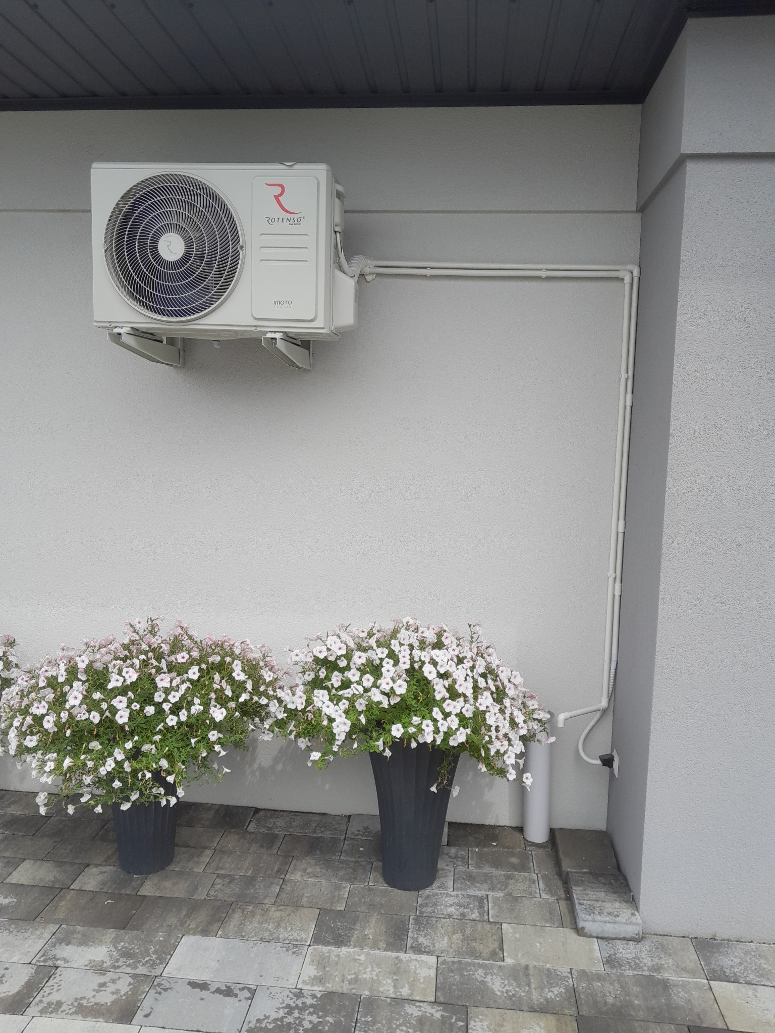 Biała jednostka zewnętrzna klimatyzatora Rotenso zamontowana na ścianie domu, z widocznymi rurami instalacyjnymi i dwoma donicami z białymi kwiatami u podstawy ściany.