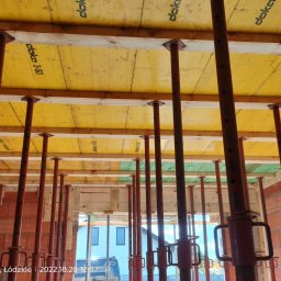 Łukasz Kwiatek - Szczegółowe ujęcie szalunku stropowego z żółtą sklejką Doka i stalowymi podporami teleskopowymi na budowie domu z czerwonej cegły.