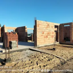 Wznoszenie ścian parterowego domu z czerwonej cegły, widoczne zbrojenia słupów i stos cegieł na fundamencie, w tle błękitne niebo.