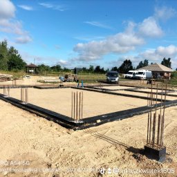 Łukasz Kwiatek - Przygotowany fundament pod dom jednorodzinny z wystającymi prętami zbrojeniowymi, zabezpieczony czarną folią izolacyjną, na piaszczystym podłożu, z widocznymi samochodami dostawczymi w tle.