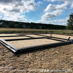 Łukasz Kwiatek - Przygotowany szalunek zbrojeniowy fundamentów na piaszczystym terenie z widocznymi wystającymi prętami zbrojeniowymi i rurami drenażowymi, w tle zabudowa jednorodzinna i drzewa.