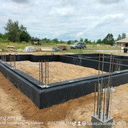 Łukasz Kwiatek - Zabezpieczone fundamenty domu jednorodzinnego z wystającymi prętami zbrojeniowymi, widok na plac budowy z domem w stanie surowym otwartym w tle.