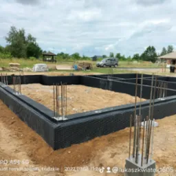 Zabezpieczone fundamenty domu jednorodzinnego z wystającymi prętami zbrojeniowymi, widok na plac budowy z domem w stanie surowym otwartym w tle.