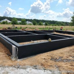 Łukasz Kwiatek - Przygotowany szalunek fundamentów domu jednorodzinnego, pomalowany na czarno, z widoczną izolacją pionową, w tle krajobraz z domami i błękitnym niebem.