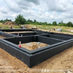 Łukasz Kwiatek - Zabezpieczone przed wilgocią fundamenty domu jednorodzinnego z hydroizolacją i folią kubełkową, widoczne zbrojenia i dwóch pracowników w tle.