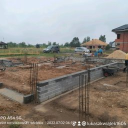 Łukasz Kwiatek - Budowa fundamentów z bloczków betonowych, zbrojenie pionowe wystające z ziemi, taczka i pracownik w kasku na budowie, w tle dom w stanie surowym otwartym i krajobraz podmiejski.