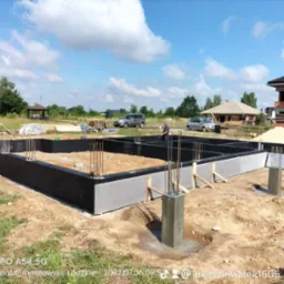 Zalane betonem fundamenty domu jednorodzinnego z wystającymi prętami zbrojeniowymi i zaizolowanymi ścianami zewnętrznymi, widok na budowę w słoneczny dzień.