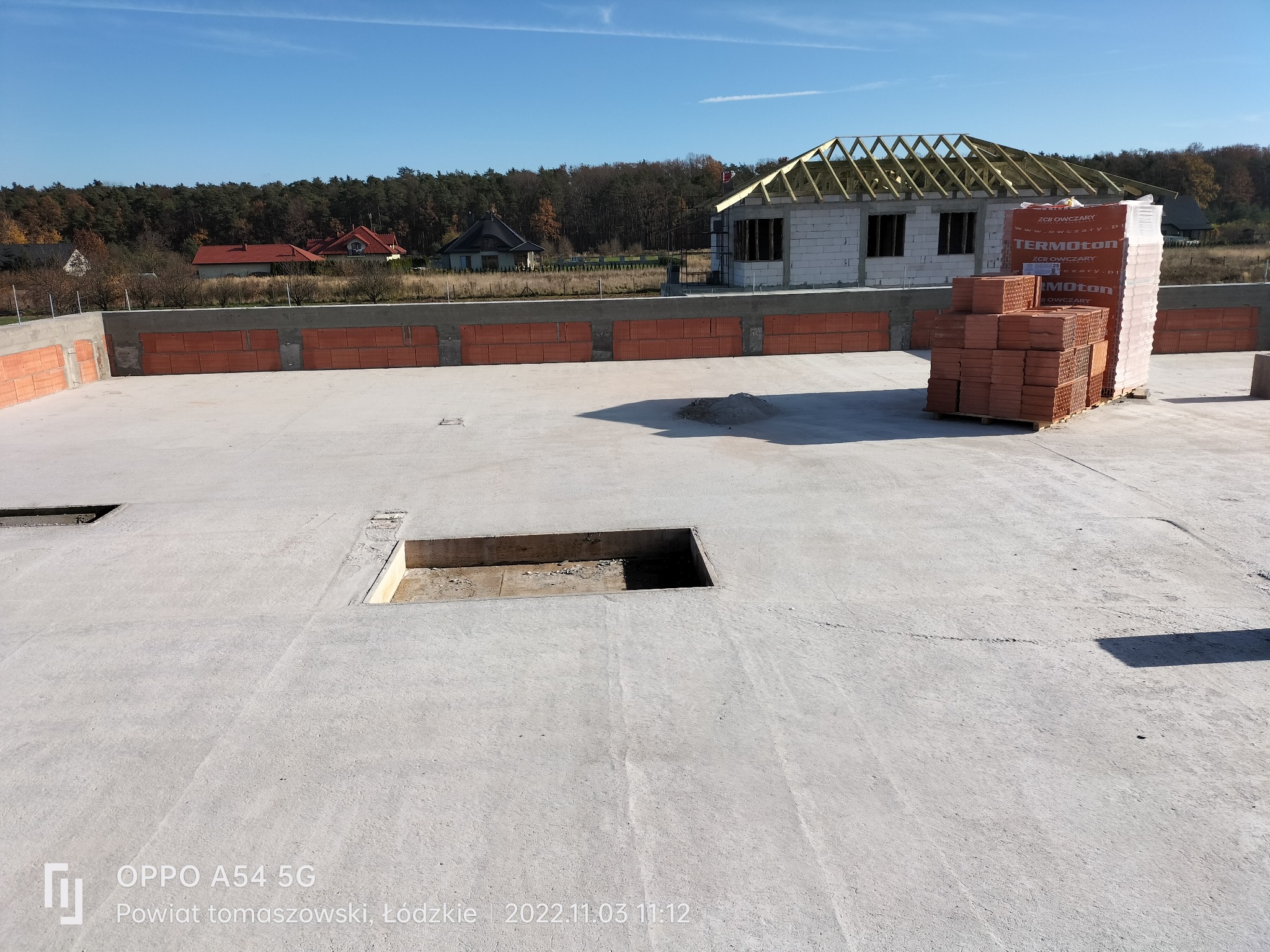 Wylana płyta fundamentowa z otworami, na której wznoszone są ściany domu z pustaków ceramicznych, w tle widoczny szkielet dachu i palety z materiałem budowlanym.