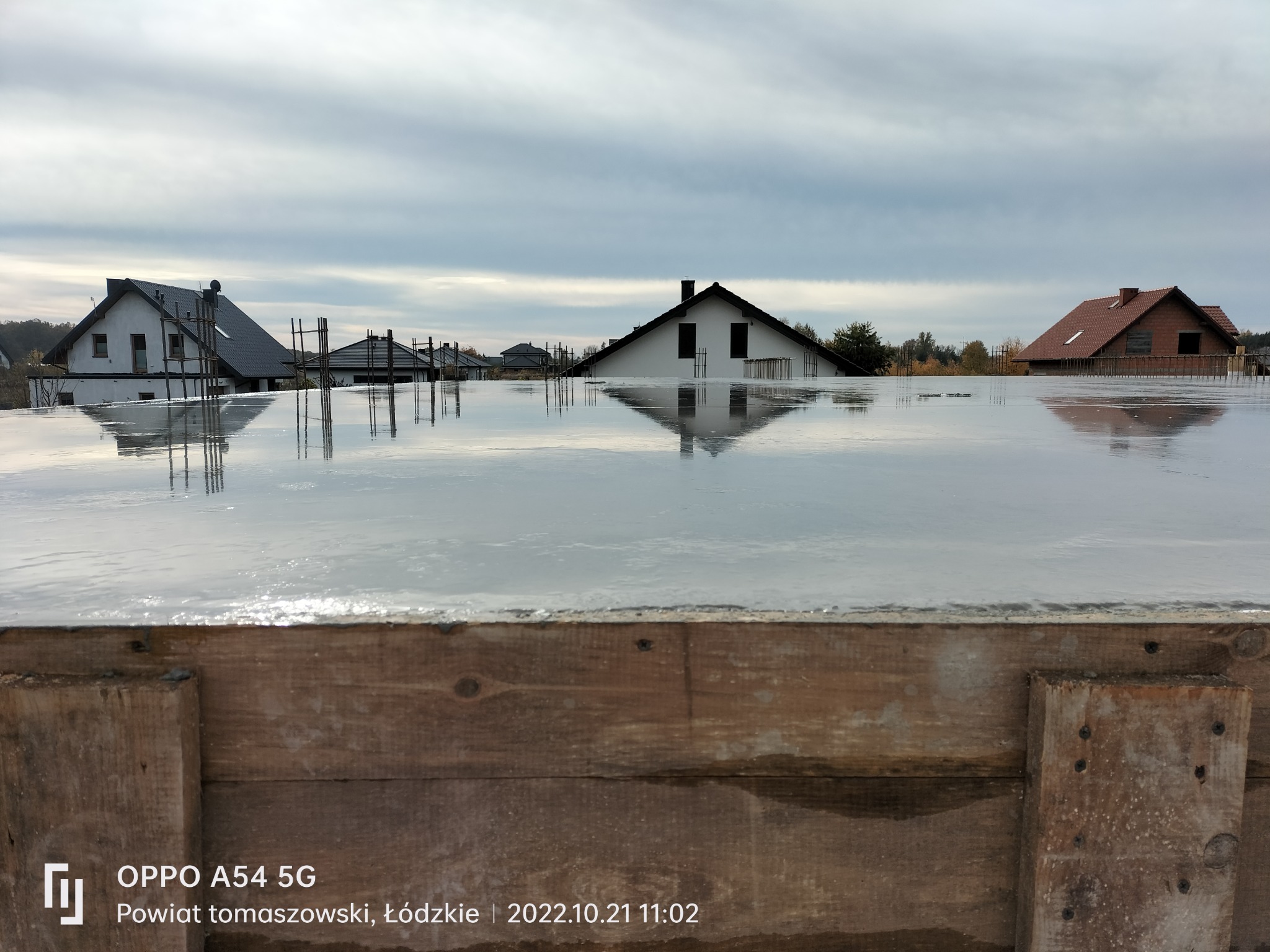 Świeżo wylany beton na fundamentach z widocznymi zbrojeniami i deskowaniem, w tle domy w stanie surowym zamkniętym, pochmurne niebo.